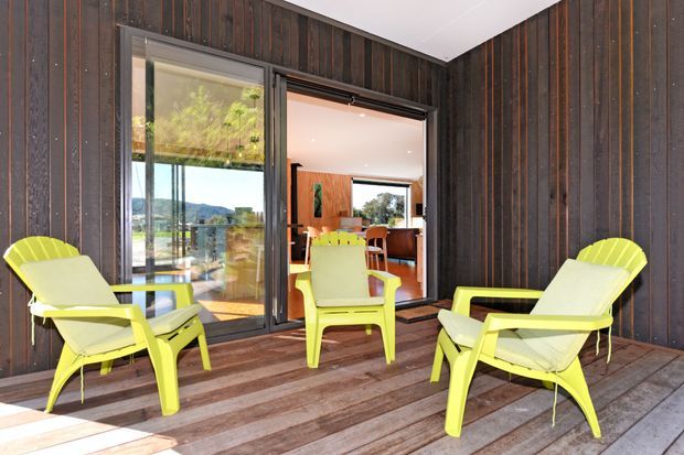 Exterior timber deck adjoining a modern home, with bright lime-green outdoor lounge chairs arranged around a central chair. Dark vertical timber cladding surrounds large glazed sliding doors, which open onto a warm timber-lined interior dining and living area. A landscaped and hilly outlook is visible through the glazing.