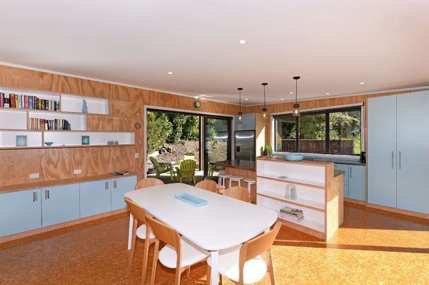 Bright open-plan kitchen and dining area with extensive plywood wall panelling, pale blue built-in cabinetry, open shelving, a large white dining table with timber chairs, pendant lighting, and garden views through glazed doors and windows. The space has a warm mid-century-inspired character with strong timber detailing and abundant natural light.