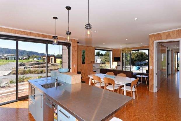 A bright, open-plan kitchen and dining area with a large stainless-steel island, timber cabinetry and wall panelling, pendant lighting, and extensive floor-to-ceiling glazing opening toward a landscaped residential setting. A dining table and living area are visible beyond the kitchen.