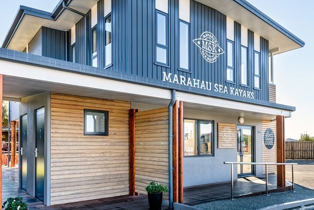 Exterior view of a contemporary two-storey commercial building for Marahau Sea Kayaks. The facade combines dark vertical metal cladding, horizontal timber weatherboards, large narrow windows, broad roof eaves, and a small accessible entry ramp with stainless-steel handrails. Signage is prominently displayed on the upper facade.