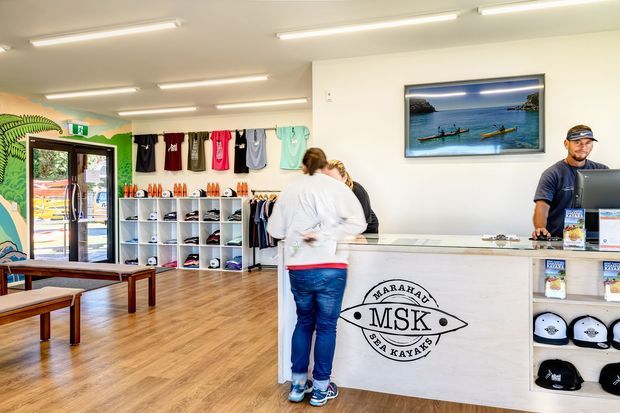 Bright retail reception and hire desk for a sea-kayaking business, with a white service counter, branded MSK Marahau Sea Kayaks graphic, shelving displaying clothing, footwear and merchandise, timber-look flooring, and framed coastal kayaking imagery. Customers and staff are present, with fluorescent ceiling lighting and a glazed entrance.