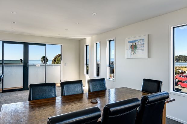 Bright contemporary dining room with a large dark timber dining table and black leather dining chairs. White walls and ceiling, recessed downlights, dark-framed glazing, and multiple windows provide expansive views toward water and a coastal residential setting. A framed artwork hangs on the wall, while the room remains clean and minimally decorated.