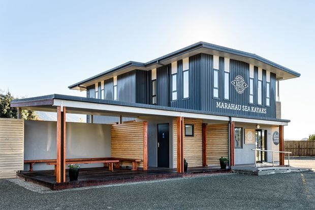 Exterior photograph of a contemporary two-storey commercial building housing Marahau Sea Kayaks. The building has a low-pitched roof with broad eaves, dark vertically ribbed metal cladding on the upper level, warm horizontal timber cladding and timber posts at ground level, extensive vertical window openings, and a covered outdoor waiting or seating area with built-in bench seating. The surrounding forecourt is gravel or dark aggregate.