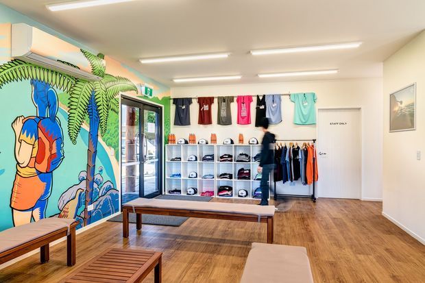 Bright, contemporary retail showroom or shop interior with a colourful illustrated mural, wall-mounted clothing displays, open shelving for caps and merchandise, a glass entry door, timber-look flooring, and simple wooden benches. Linear fluorescent-style ceiling lights provide even illumination.