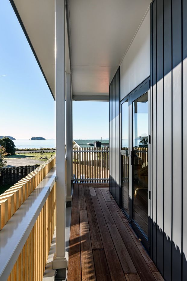 A narrow elevated timber balcony or covered terrace running alongside a contemporary coastal home. The space features dark timber decking, pale timber balustrades, white structural posts and soffit, and dark vertical metal wall cladding with large glazed doors. A bright ocean and coastal landscape are visible beyond the railing.