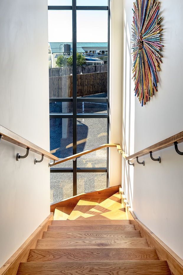 Bright interior stairway descending toward a large floor-to-ceiling black-framed glazed window. The staircase has warm timber treads and trim, white walls, timber handrails supported by dark metal brackets, and a colourful linear wall artwork on the right. Strong sunlight creates geometric shadows across the landing and lower steps, with an exterior residential setting visible through the glazing.