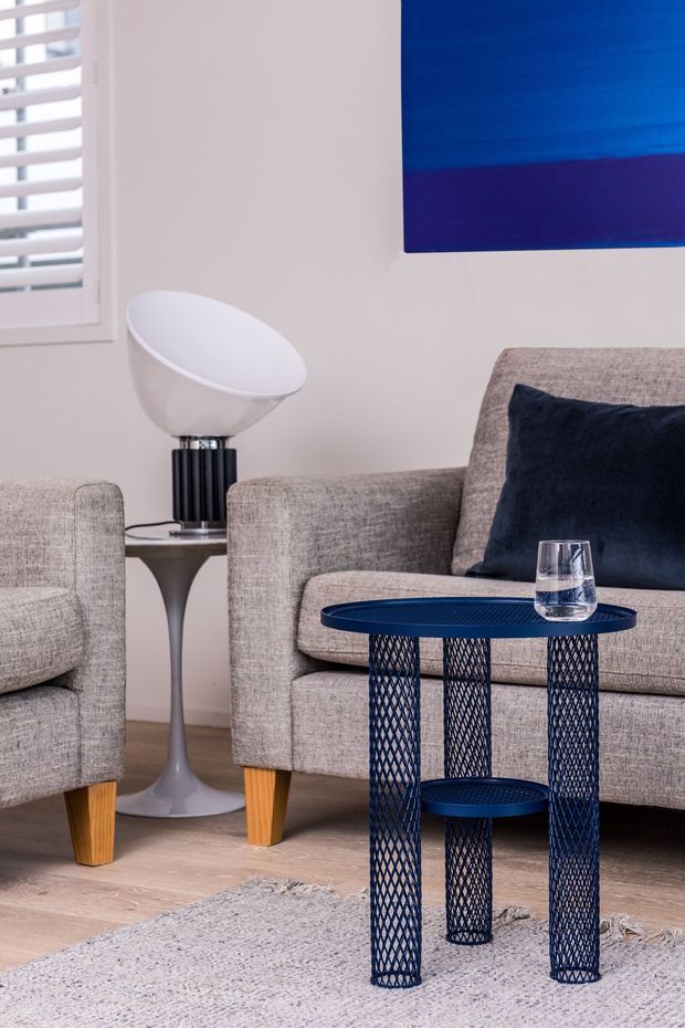 A close-up of a refined living room seating area with two textured taupe-grey upholstered armchairs and sofa, warm timber legs, a sculptural side table and modern bowl-shaped lamp. A dark navy geometric metal coffee table and navy cushion provide contrast, while a blue abstract artwork, pale timber flooring, and a light textured rug complete the space.