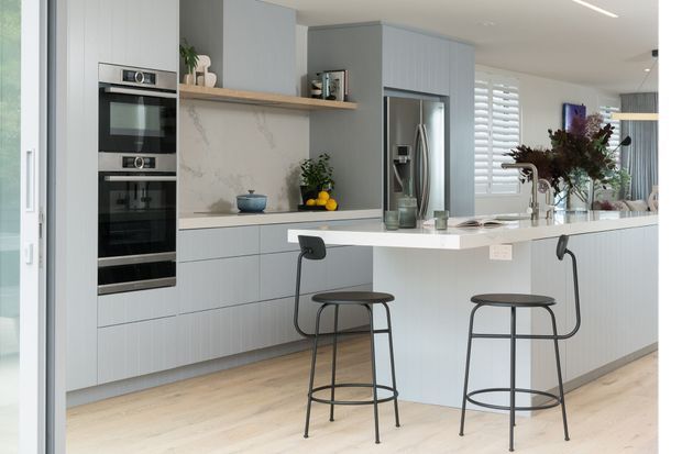 Bright contemporary kitchen with pale grey flat-panel cabinetry, integrated double ovens, a large central island with a white benchtop, two black bar stools, stainless-steel refrigerator, marble-look splashback, timber shelving, and light timber flooring. The space opens toward a glazed area with plantation shutters and abundant natural light.