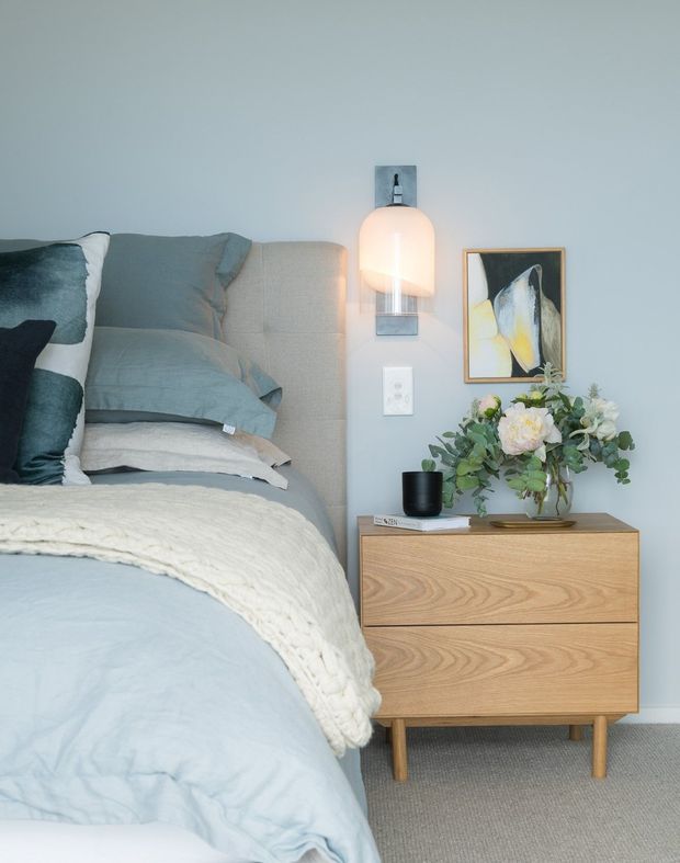 A close-up of a softly styled bedroom corner featuring a pale blue upholstered bed with layered blue-grey bedding and a textured cream throw. Beside it is a light timber two-drawer bedside cabinet with tapered legs, topped with a black vessel and floral arrangement. A wall-mounted contemporary light, framed abstract artwork, and pale blue-grey wall complete the calm, understated scheme.