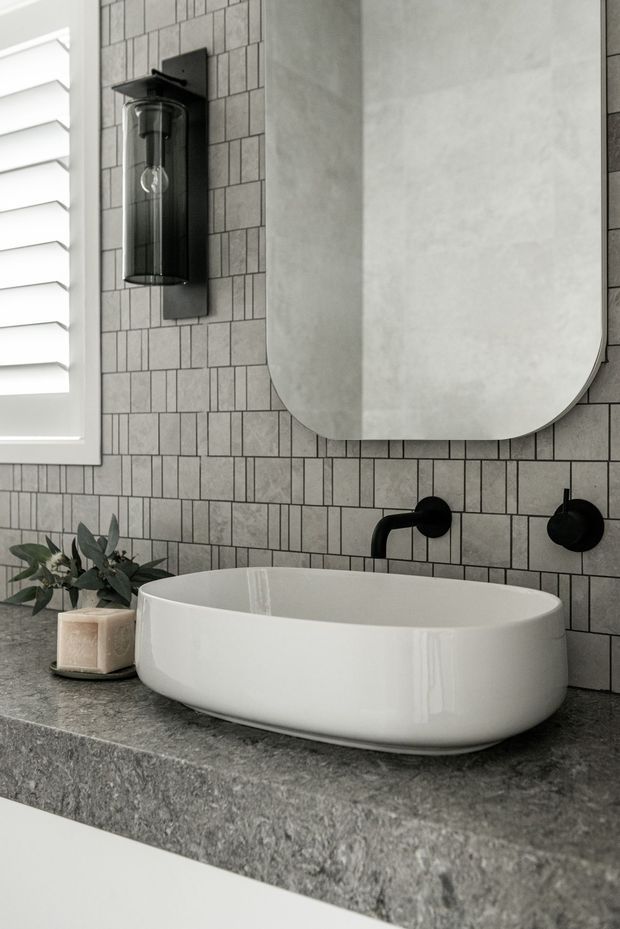 Close-up of a refined bathroom vanity featuring a white vessel basin on a dark grey natural-stone benchtop, with matte black wall-mounted tapware, a rounded rectangular mirror, grey rectangular tiled wall, black wall sconce, and white plantation shutters.