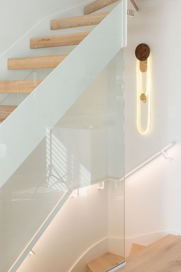 Bright contemporary stairway with pale timber floating treads, a clear glass balustrade, and a white structural stair stringer. A warm vertical wall light with timber accents adds a subtle feature detail, while light timber flooring continues at the lower level.