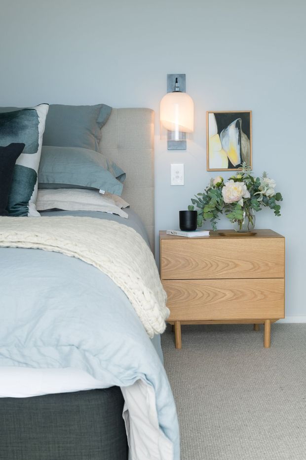 A softly styled bedroom corner with a low upholstered bed, layered pale blue and white bedding, and a warm timber two-drawer bedside table. The light blue-grey wall features a wall-mounted pendant light and framed abstract artwork, while a vase of greenery and flowers adds a natural accent. The room has a calm, refined, contemporary feel with carpeted flooring.
