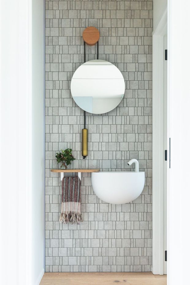 A compact bathroom or powder room viewed straight on through a doorway, featuring a wall-mounted white vessel basin, a round mirror suspended from a black strap, a brass pendant light, a narrow timber shelf, and a full-height grey patterned tile wall. A small plant and hanging hand towel add warmth, while pale timber flooring and white trim frame the space.