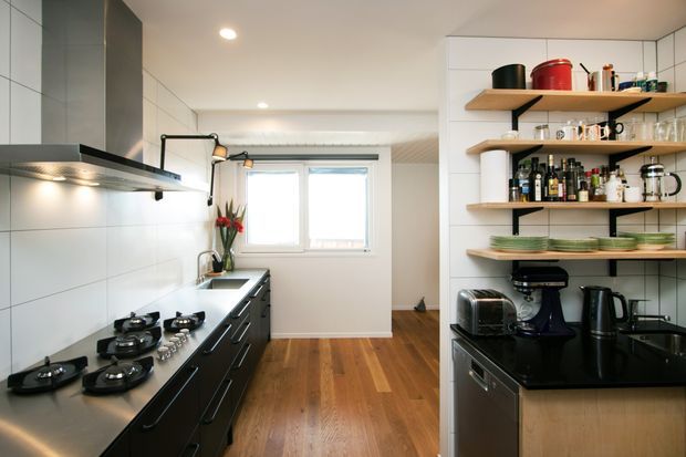 A contemporary galley-style kitchen with black cabinetry and benchtops, stainless-steel appliances, white tiled walls, open timber shelving, and warm timber flooring. The kitchen includes a long cooktop run, rangehood, sink area, coffee appliances, and a bright window at the far end.