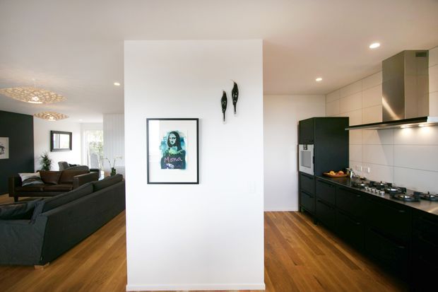 Open-plan contemporary kitchen and living area with warm timber flooring, matte black kitchen cabinetry, white tiled splashback, and a central white partition wall displaying artwork. A dark sectional sofa and pendant lighting are visible in the living zone.