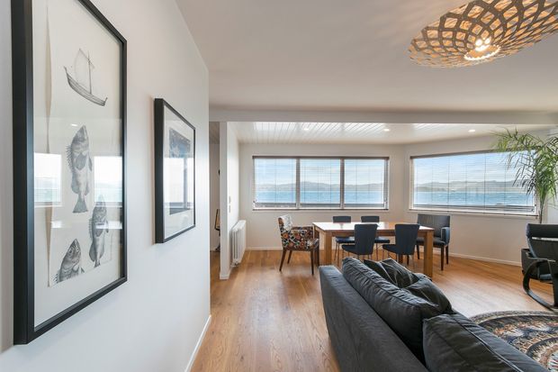 Bright open-plan living and dining area with polished timber flooring, white walls, framed marine artwork, a dark leather sectional sofa, and a timber dining table with mixed seating. Large windows provide expansive water views, while a woven sculptural pendant light adds a natural coastal accent.