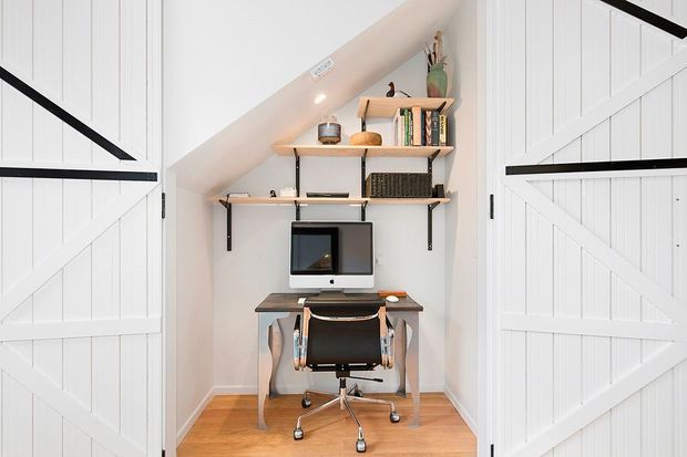 A compact home office nook tucked beneath a sloping ceiling and enclosed by open white barn-style doors. The space features a small wooden desk, black office chair, desktop computer, floating timber shelves with books and decor, white vertical wall panelling, black metal shelf brackets, and warm timber flooring.