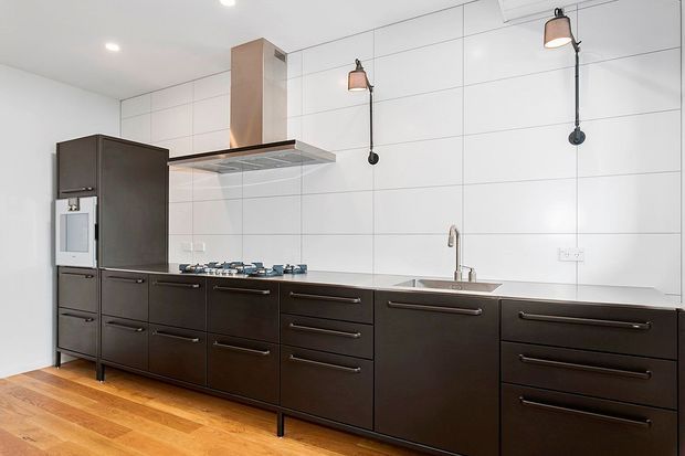 Contemporary galley-style kitchen with a long run of dark cabinetry, integrated appliances, stainless-steel cooktop and rangehood, white tiled walls, and a timber floor. Black wall-mounted pendant lights and a stainless-steel sink and tap add industrial accents.