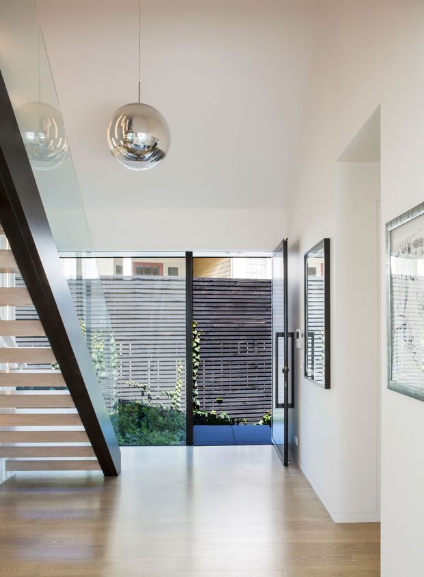 Bright contemporary entry hall with a floating timber staircase, dark structural stringer, glass balustrade, pale timber flooring, and white walls. Chrome spherical pendant lights hang from the high ceiling. Full-height glazed doors open to a small planted courtyard screened by horizontal timber slats, with framed artwork on the right wall.