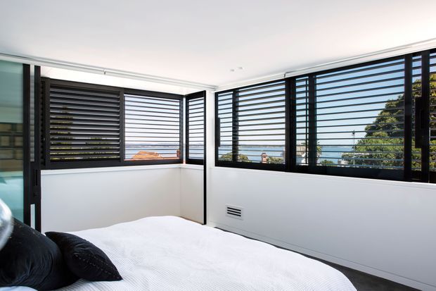 Bright modern bedroom with a white bed and dark bedding, enclosed by large black-framed horizontal louvred windows. The windows provide expansive views toward blue water and surrounding greenery, while white walls and ceiling create a clean, airy interior.