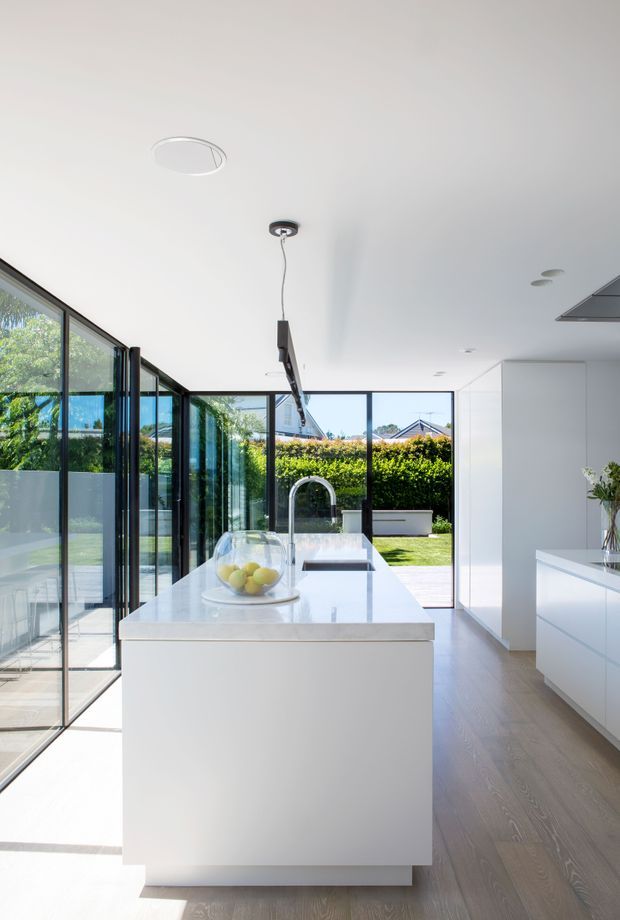 Bright, minimalist contemporary kitchen with a large white waterfall-edge island, integrated cooktop, white cabinetry, and pale timber flooring. Full-height black-framed glazing opens the room toward a green backyard, while a slim linear pendant hangs above the island. The space is clean, airy, and highly connected to the outdoors.