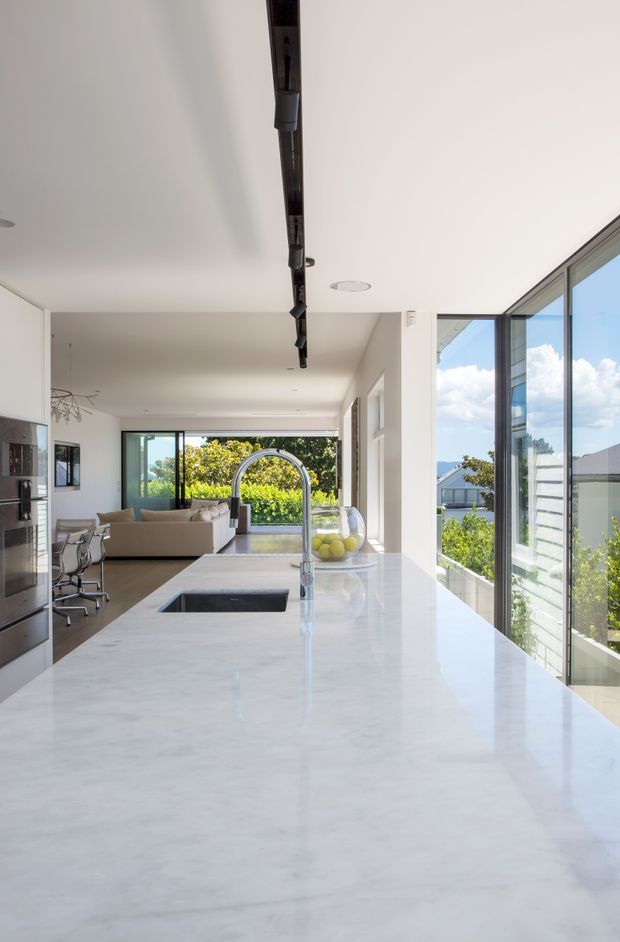 A bright, open-plan contemporary kitchen viewed along a large white marble island with an undermount sink and tall curved chrome mixer tap. White fixed cabinetry and integrated appliances line the left wall, while full-height glazing on the right opens to leafy suburban views. A living area with a neutral sofa is visible beyond the kitchen, and a dark linear ceiling track feature runs overhead.