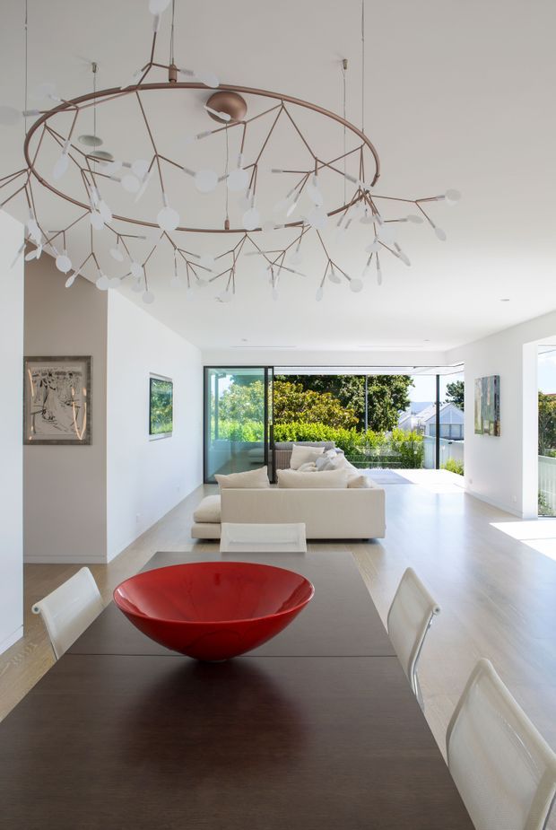 Bright open-plan interior viewed from the dining area toward a contemporary living room. A long dark timber dining table with white chairs and a vivid red bowl occupies the foreground, while a cream sectional sofa sits beyond. Floor-to-ceiling sliding glass doors open to a leafy outdoor setting, and a large sculptural branch-like pendant light spans the ceiling. White walls, pale flooring, abundant daylight, and clean modern lines create an airy, refined space.