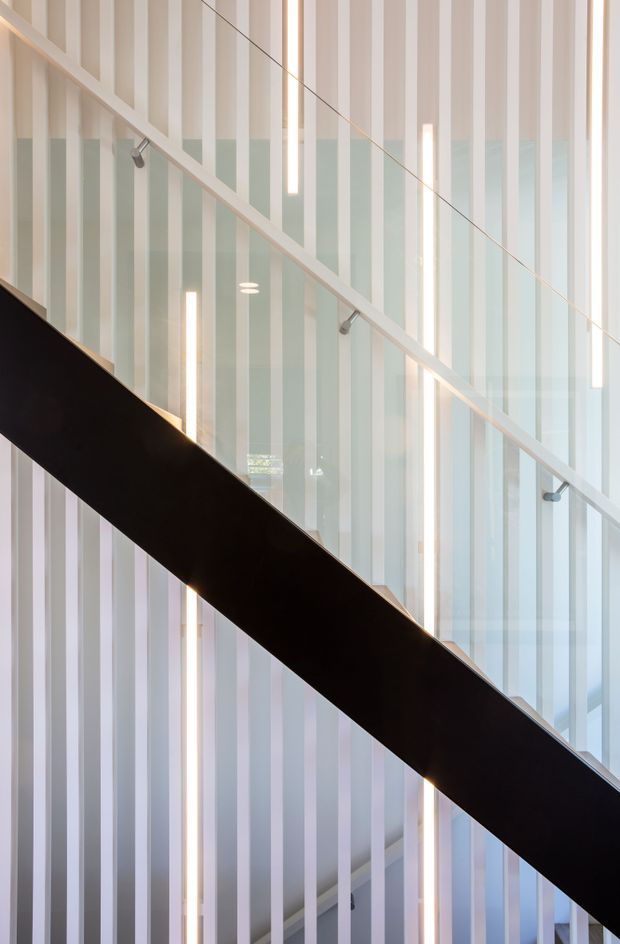 Abstract architectural interior view of a modern stairway. A bold dark diagonal stair flight crosses in front of a tall translucent vertical slatted screen, with a slim pale handrail, glass elements, and repeated linear pendant or integrated lights creating a strong geometric composition.