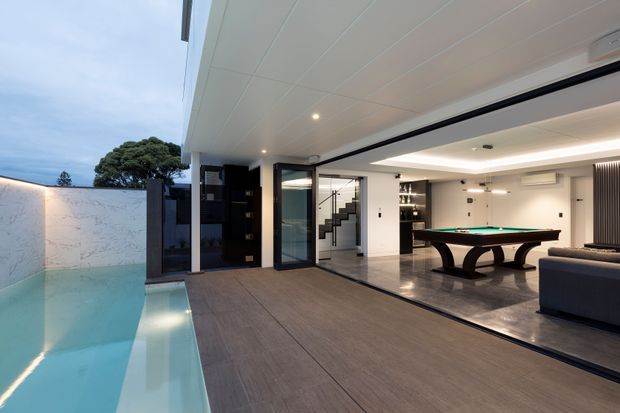 Contemporary covered outdoor entertaining area opening directly into a polished games and living space, with a narrow swimming pool along the left, dark timber-look decking, recessed ceiling lighting, black-framed glazing, and a visible billiards table indoors. A staircase and small bar or display shelving are visible in the background.