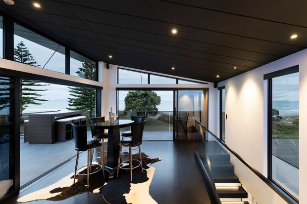 Contemporary open-plan interior with a compact bar-height dining setting beside extensive floor-to-ceiling glazing. The dark ceiling, polished dark floor, white walls, recessed downlights, and black-framed windows create a refined modern look. A stair opening with illuminated steps leads downward, while an outdoor terrace, coastal vegetation, and water are visible through the glazing.