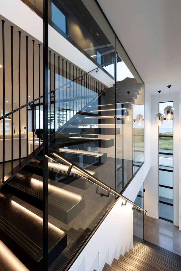 A dramatic modern interior stairway with multiple flights of open-riser stairs, dark timber treads, black metal balustrades, extensive glass panels, and integrated linear lighting. The stairwell rises through a bright double-height space with white walls, large windows, pendant lights, and glimpses of adjoining living areas.
