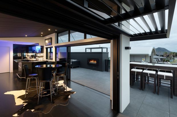 A sophisticated indoor-outdoor entertaining area with a dark contemporary kitchen and home bar, a raised island with bar stools, and large sliding glass doors opening to a covered terrace. The terrace includes a built-in outdoor bar, lounge seating, and a wall-mounted fireplace, with a semi-urban landscape visible beyond. Strong black, charcoal, timber, and cool grey finishes create a dramatic evening atmosphere.