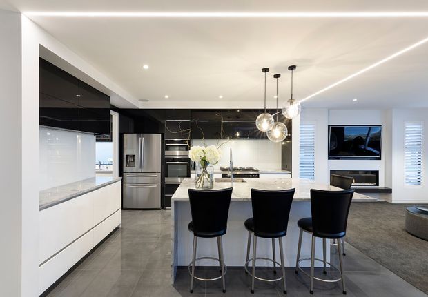 A contemporary open-plan kitchen and living area viewed from the kitchen. The kitchen features a large white island with three black bar stools, white handleless cabinetry, a dark glossy feature wall with subtle veining, stainless-steel appliances, and three spherical pendant lights. Grey tiled flooring extends toward a carpeted living area with built-in media joinery and plantation shutters. Recessed ceiling lighting and linear LED lighting create a clean, polished appearance.