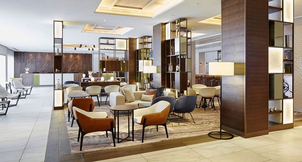 A sophisticated contemporary hotel or commercial lounge with multiple seating areas, dark timber room dividers and shelving, warm wood-look flooring, pale stone or tile flooring, layered ceiling lighting, and floor-to-ceiling glazing. The space is open, bright, and arranged as a refined reception or lobby lounge.