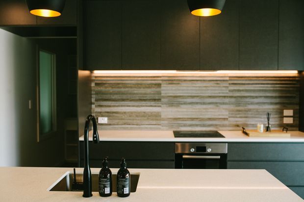 A contemporary kitchen viewed toward a dark, streamlined cabinetry wall. The space features a large pale stone island with integrated sink and black gooseneck tap, a built-in oven and cooktop, dark cabinetry, a horizontally textured stone or tile splashback, and warm pendant lighting.