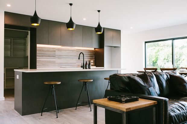 Open-plan contemporary kitchen and living area with dark flat-panel cabinetry, a large island with seating, wood-look backsplash, and three black pendant lights. A black leather sofa and timber coffee table occupy the foreground, while a large window provides abundant natural light.