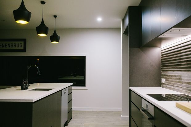 Contemporary galley-style kitchen with a central island, dark cabinetry, white benchtops, integrated appliances, and a textured horizontal tile splashback. Black pendant lights hang over the island, while a large dark-framed window adds contrast to the otherwise neutral space.