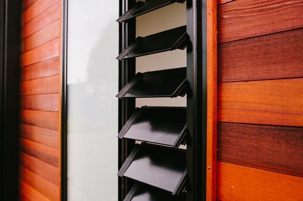 Close-up exterior detail of a black louvred window set within warm reddish-brown horizontal timber cladding. The adjustable louvres are partially open, contrasting with the smooth dark timber boards and dark window framing.