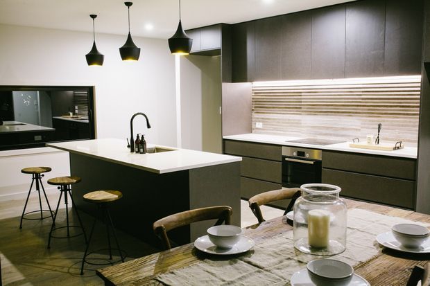 Contemporary open-plan kitchen and dining area with a large dark island, white benchtop, integrated dark cabinetry, horizontal timber-look splashback, pendant lighting, and a rustic timber dining table with stools and chairs.
