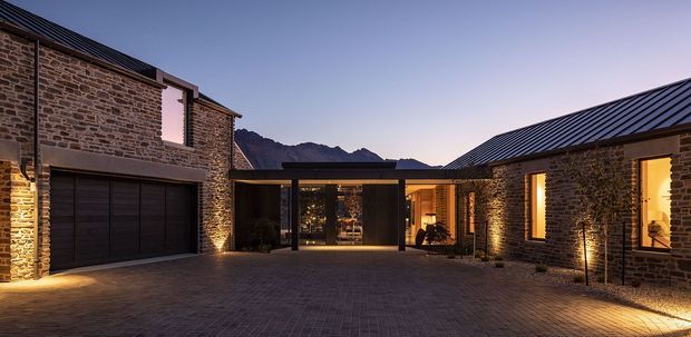 Twilight exterior view of a low-rise, courtyard-style residence composed of dark natural stone masonry wings linked by a glazed central entry or living volume. Dark standing-seam pitched roofs, large dark-framed openings, integrated garage doors, warm architectural lighting, and a paved brick courtyard define the scene, with rugged mountains visible beyond.