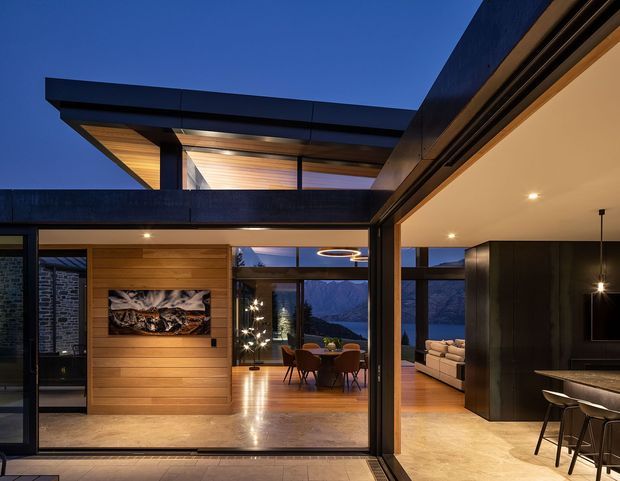 Twilight view into a sophisticated open-plan modern home, with dark metal-framed glazing and roof forms, warm timber wall and ceiling lining, polished timber floors, a dining area, living room, and kitchen with island seating. Large windows frame a mountainous landscape, while recessed and pendant lighting create a warm evening atmosphere.