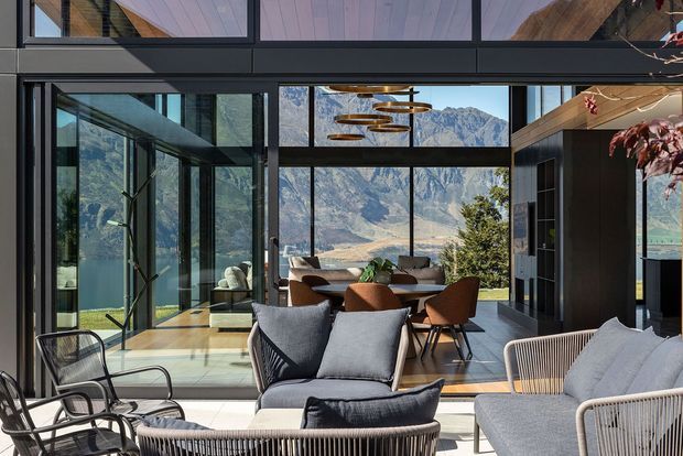 A bright contemporary open-plan living and dining area with floor-to-ceiling black-framed glazing overlooking dramatic alpine mountains. The room features a large light-grey sectional sofa, woven lounge chairs, a timber dining table with sculptural brown chairs, dark built-in cabinetry, and layered circular pendant lights. Timber flooring and warm timber ceiling accents add natural warmth to the otherwise crisp, modern interior.