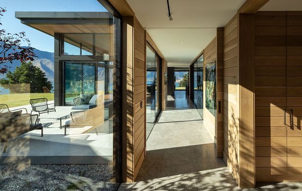 A bright modern residential entry hall viewed from the threshold, with extensive floor-to-ceiling glazing opening toward a furnished outdoor terrace and mountain landscape. The corridor features warm horizontal timber wall cladding and built-in timber cabinetry, pale polished stone or concrete flooring, slim dark window and door frames, and a clean flat ceiling with discreet linear lighting. Strong indoor-outdoor connection and abundant natural light define the space.