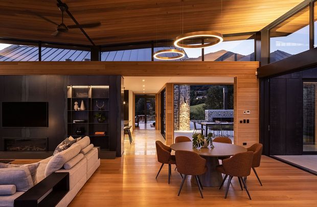 A warm, contemporary open-plan living and dining area with extensive timber ceilings and flooring, dark built-in media cabinetry, a large upholstered sectional sofa, and a round dining table with upholstered chairs. Floor-to-ceiling glazing opens views toward a mountainous landscape and a stone-clad outdoor area. Circular pendant lights and integrated lighting add a refined modern character.