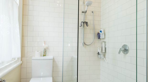 Bright, compact contemporary bathroom with white subway-tile walls, a glass-enclosed walk-in shower, chrome shower fixtures, and a white toilet. Natural light enters through the curtained window, creating a clean and airy appearance.