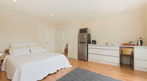 Bright, compact bedroom with a white bed, pale walls, timber flooring, built-in white cabinetry and drawers, a small desk, and an integrated kitchenette area with a refrigerator and countertop appliances. The room has recessed ceiling lights and a clean, simple contemporary fit-out.