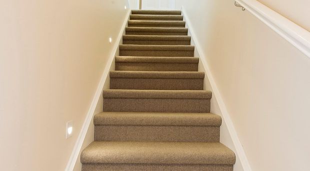A narrow residential staircase viewed from the bottom, with fully carpeted brown-beige treads and risers, white painted walls, white trim, recessed wall lights, and a simple white handrail. The stairway leads upward toward a bright landing.