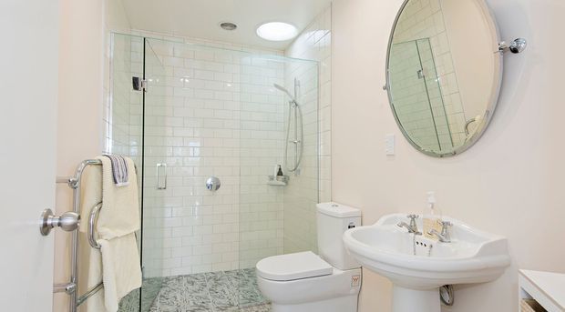 Bright compact bathroom viewed from the doorway, featuring a frameless glass walk-in shower with white subway-tile walls, patterned grey floor tiles, a toilet, pedestal sink, round wall mirror, and chrome fixtures. A towel rail is mounted near the shower entrance.