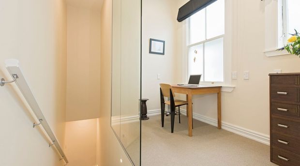 Bright compact home office nook beside a stairwell, featuring a simple timber desk and chair, laptop, large window with roller blind, beige carpet, white walls with decorative skirting, and a dark timber chest of drawers. A glass stair balustrade and handrail are visible at left.