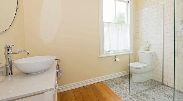 Bright traditional bathroom featuring a freestanding vessel basin on a light vanity, chrome fixtures, a white toilet, and a frameless glass shower enclosure. The shower has white subway-tile walls and patterned floor tiles, while the main room has warm timber flooring, cream walls, and decorative skirting.